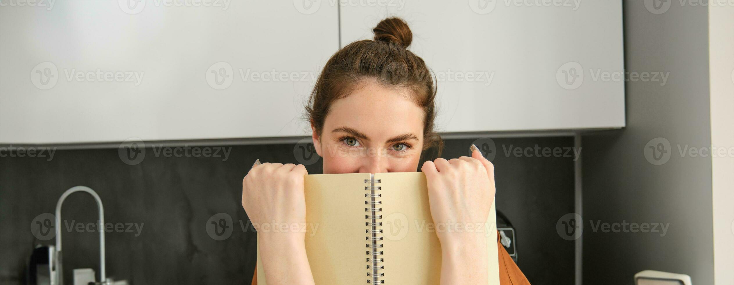 Portrait of smiling woman hiding her smile behind notebook, giggling and looking at camera flirty, standing in the kitchen with notes photo