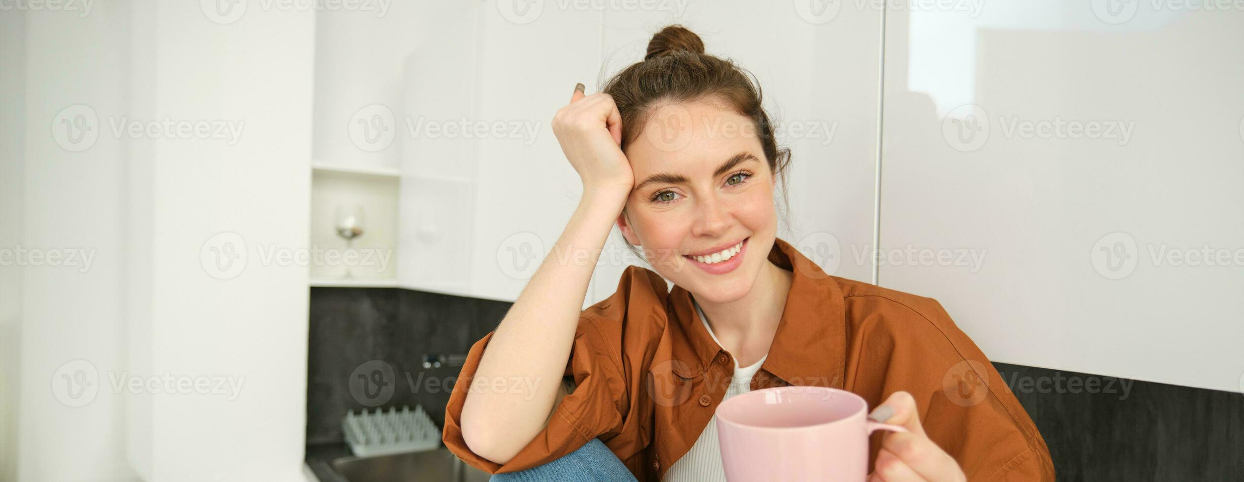 Portrait of woman with cup of coffee in hand, sits in kitchen and smiles at camera photo