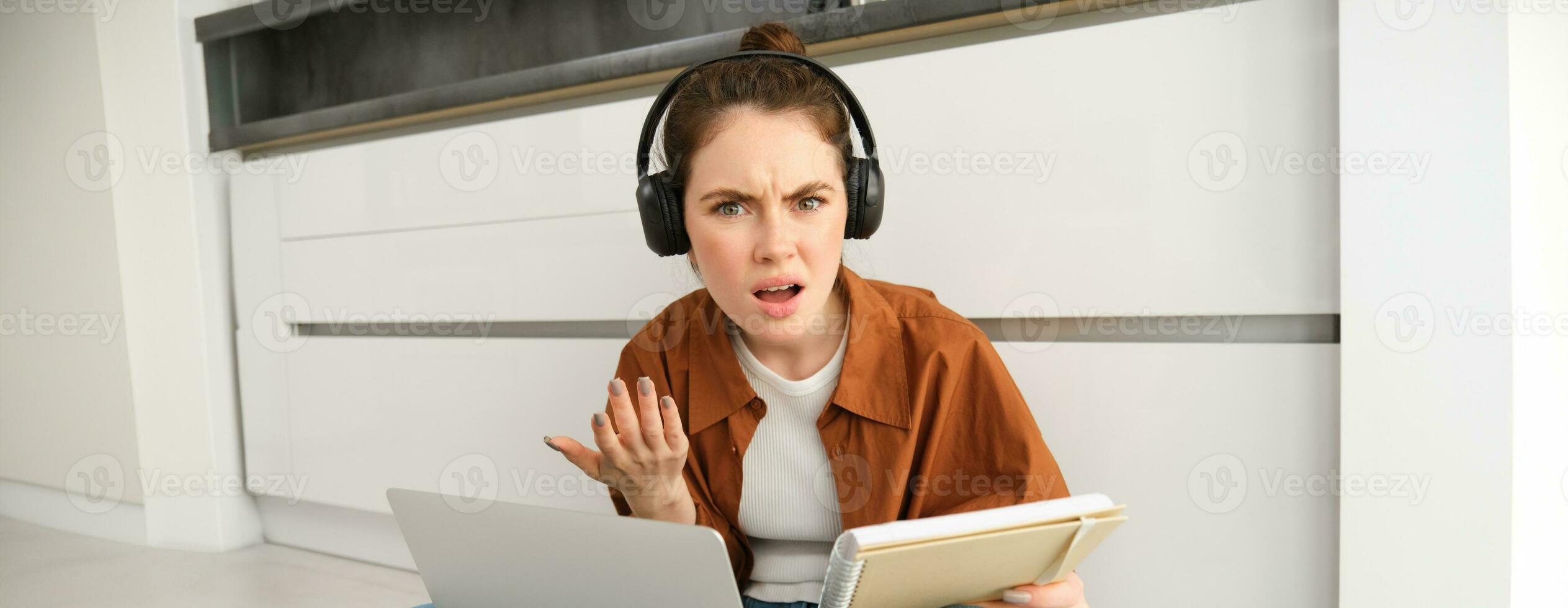 Portrait of annoyed and confused woman in headphones, sits on floor with laptop and notebook, looks frustrated, has issues in doing homework or studying photo