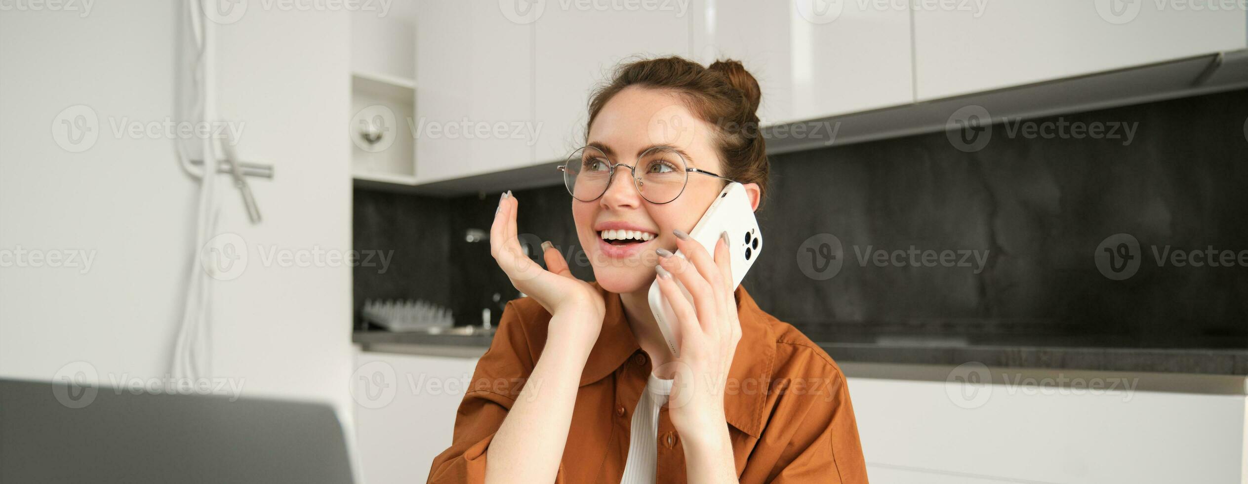 retrato de joven mujer de negocios, Trabajadores por cuenta propia dama trabajando desde hogar, haciendo teléfono llamadas a clientela y sonriente, utilizando ordenador portátil en cocina foto