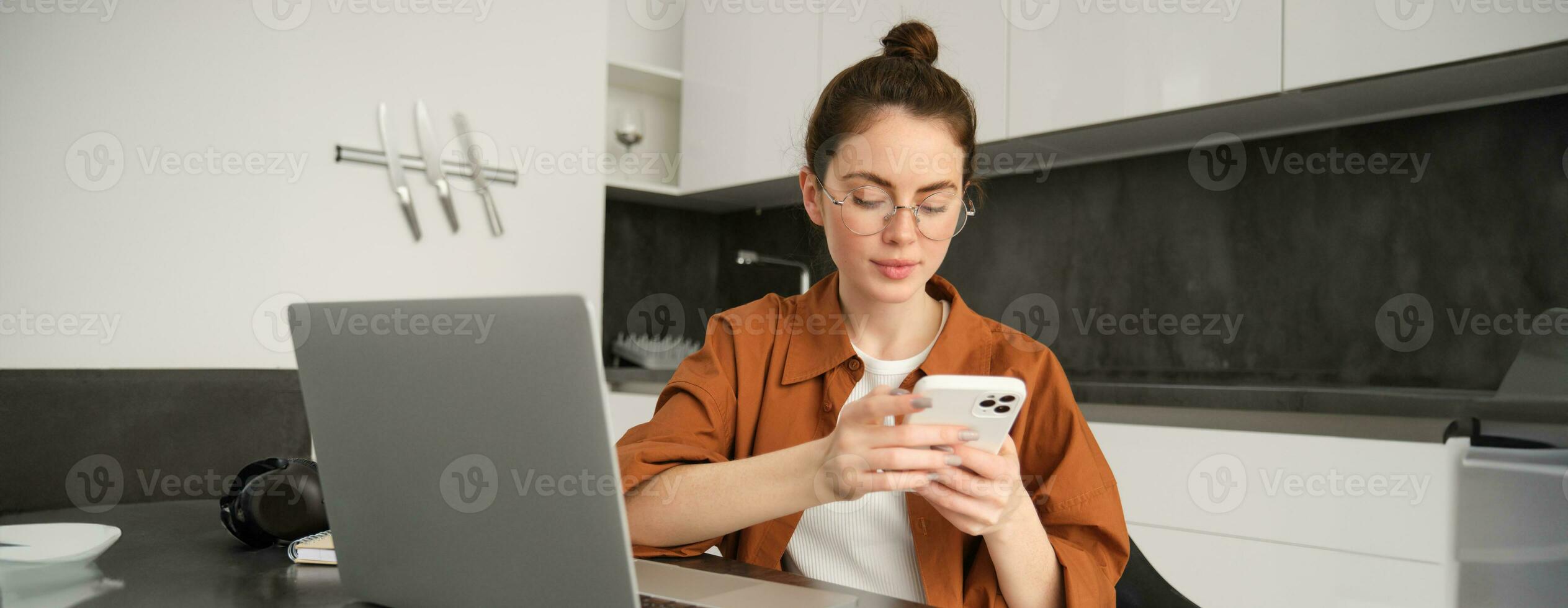 Portrait of young female freelancer, woman working from home with laptop, girl programmer sitting with computer in kitchen, answer message on smartphone photo