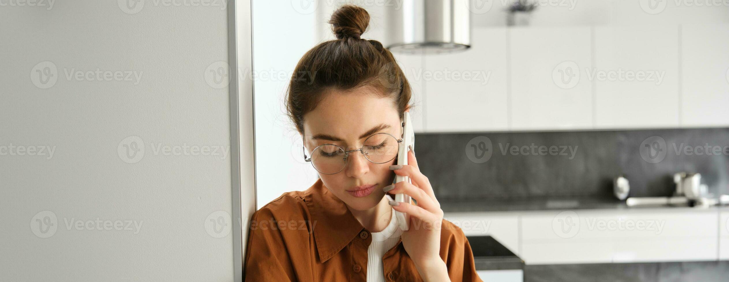 retrato de joven triste mujer en anteojos, hablando en móvil teléfono, escuchando a voz mensaje en teléfono inteligente con decepcionado, preocupado cara expresión, en pie a hogar, propensión en pared foto