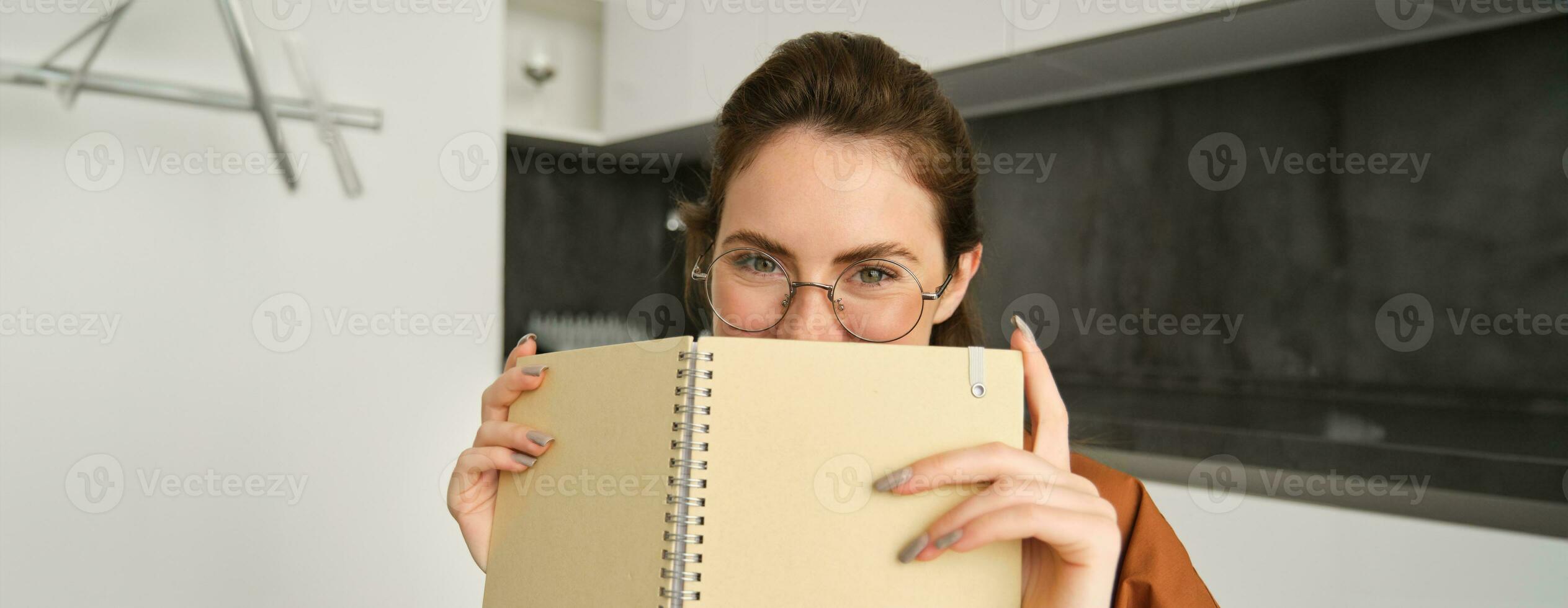 Close up portrait of cute brunette woman in glasses, sitting at home, smiling, hiding face behind yellow notebook photo