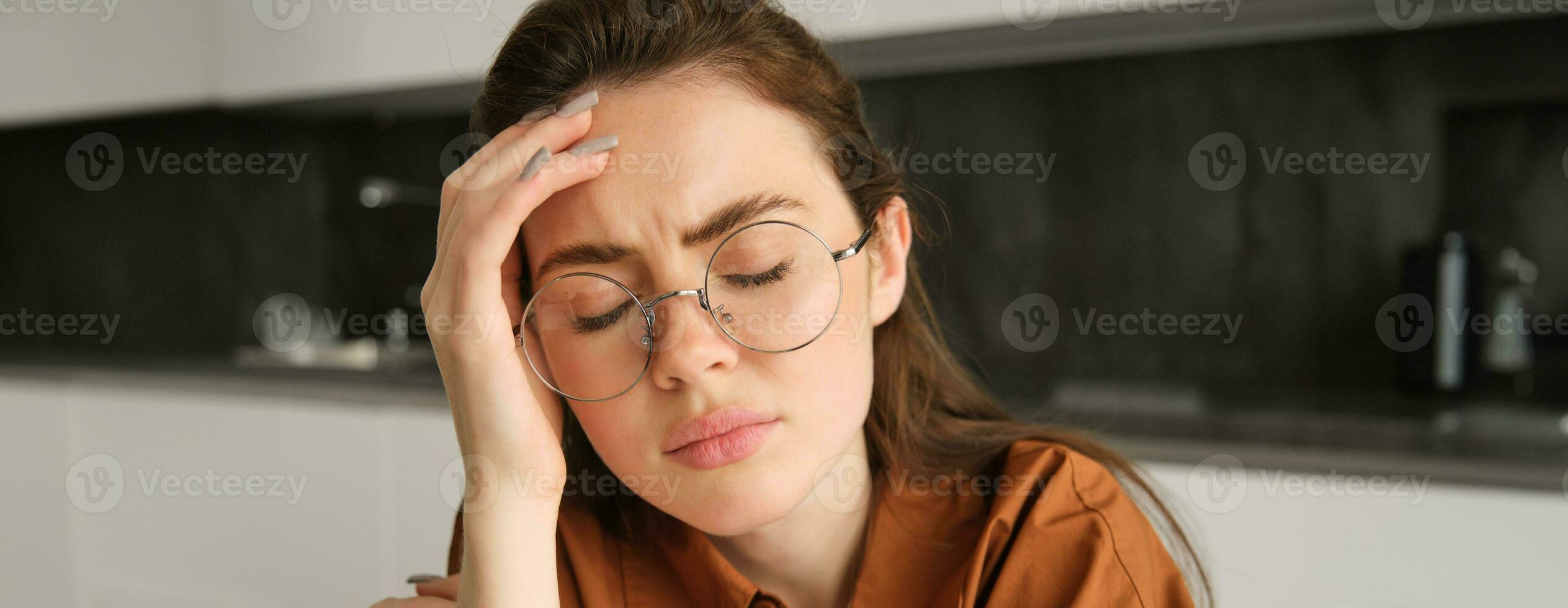 Portrait of tired young working woman in glasses, holds hands on head, suffers painful migraine, has pain in temples, sitting with headache in kitchen photo