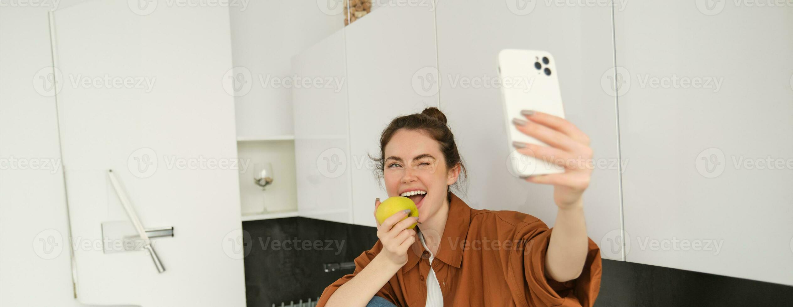 Beautiful young woman, lifestyle blogger records video of herself, takes selfie while biting an apple, sitting on kitchen counter, using smartphone photo