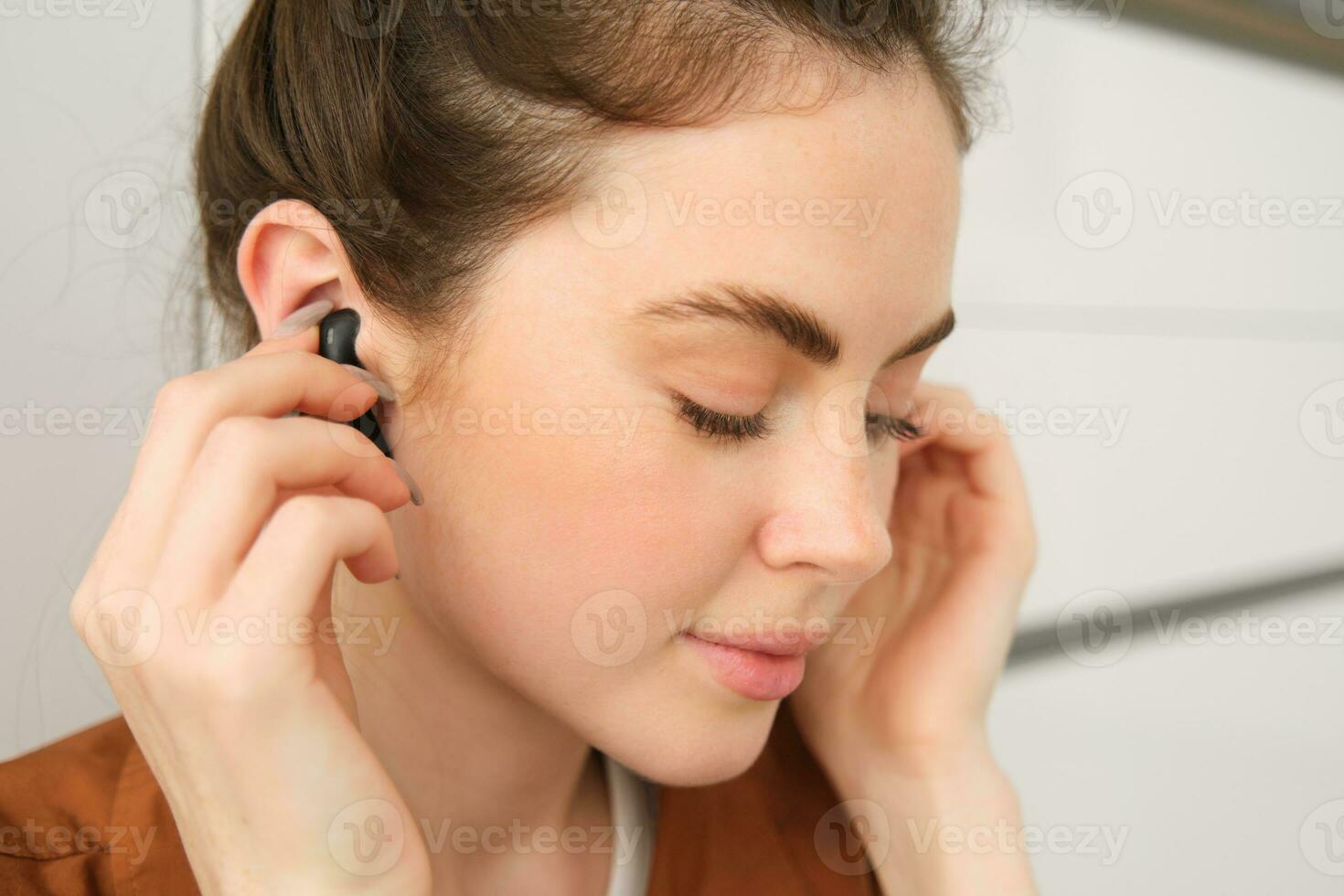 Close up portrait of tender, smiling and cute woman, puts on black wireless earphones in her ears, listens to music photo