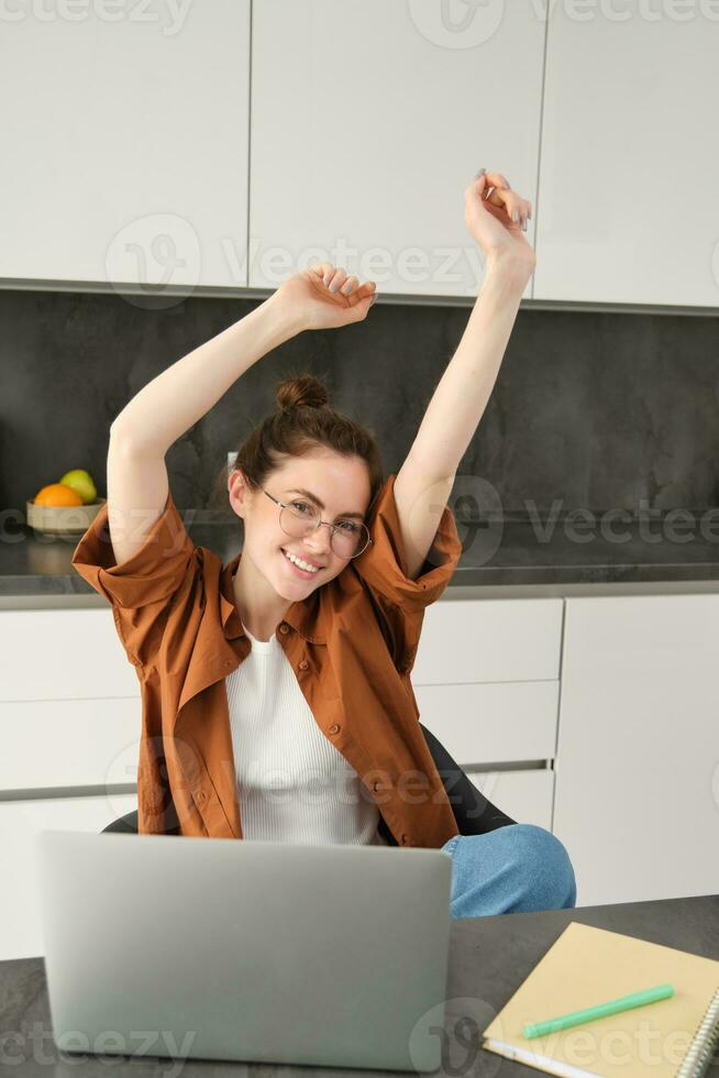 Vertical shot of smiling, lively young woman in glasses, working on remote, sitting at home with laptop, using computer to study online, stretching hands with pleased emotion photo