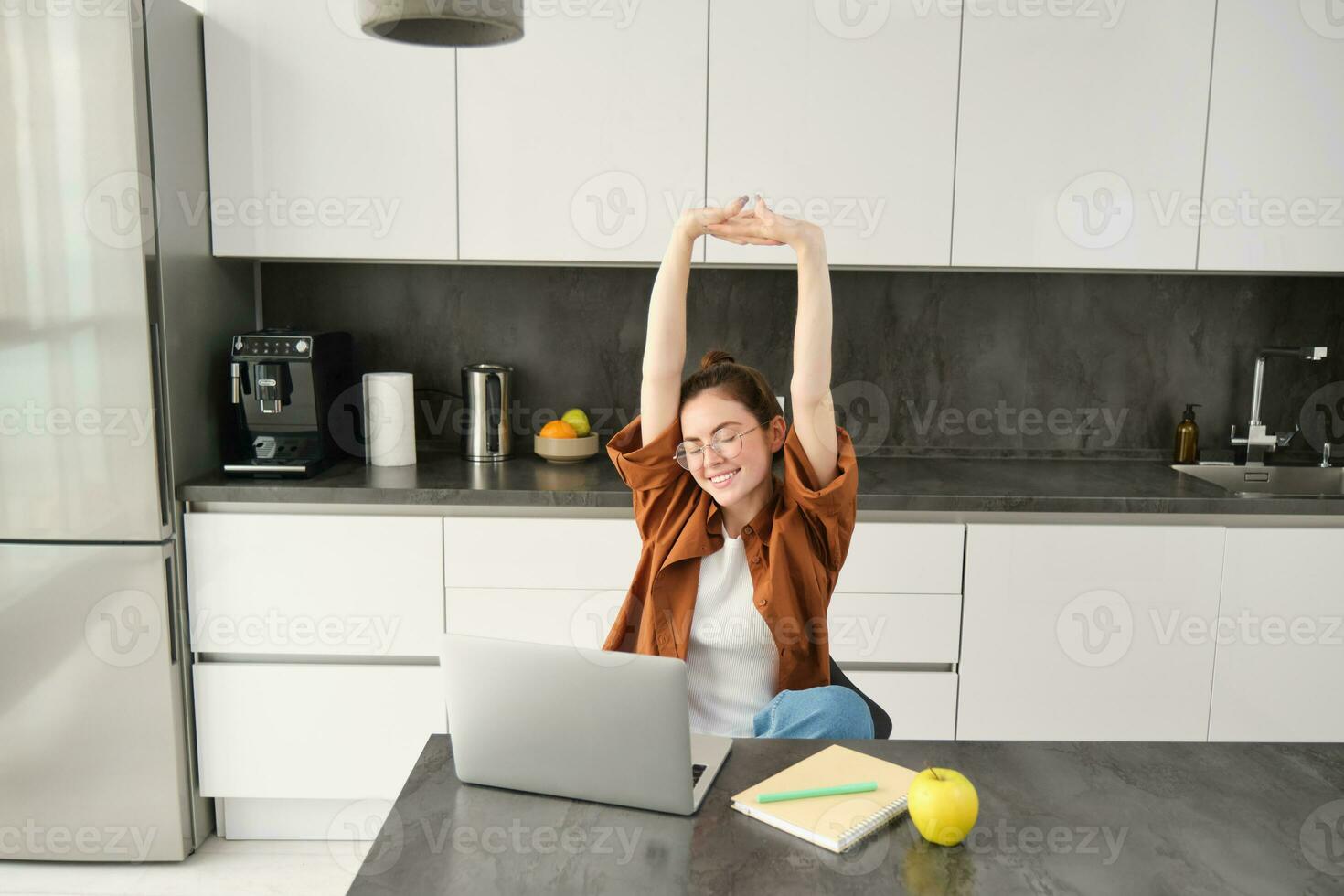 Productivity and lifestyle concept. Beautiful young woman finishes working, stretching hands with satisfied expression, sitting in kitchen with laptop and documents, has remote workplace at home photo