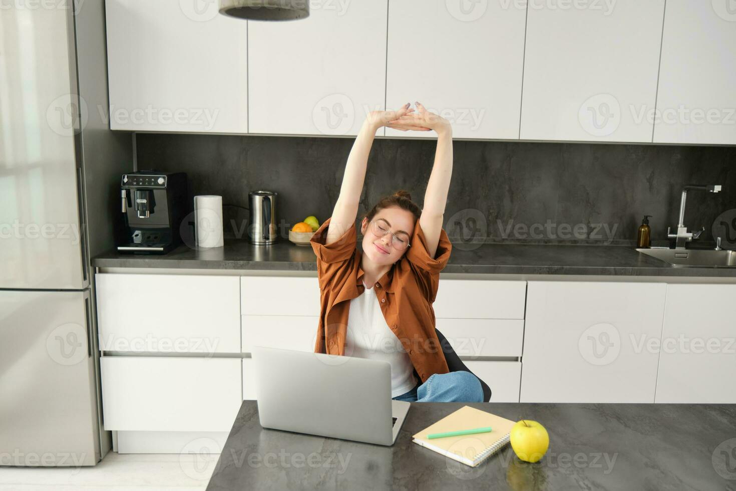 Portrait of smiling, pleased woman in glasses, sitting in kitchen, finish working on project, using laptop, stretching hands with satisfied face expression photo