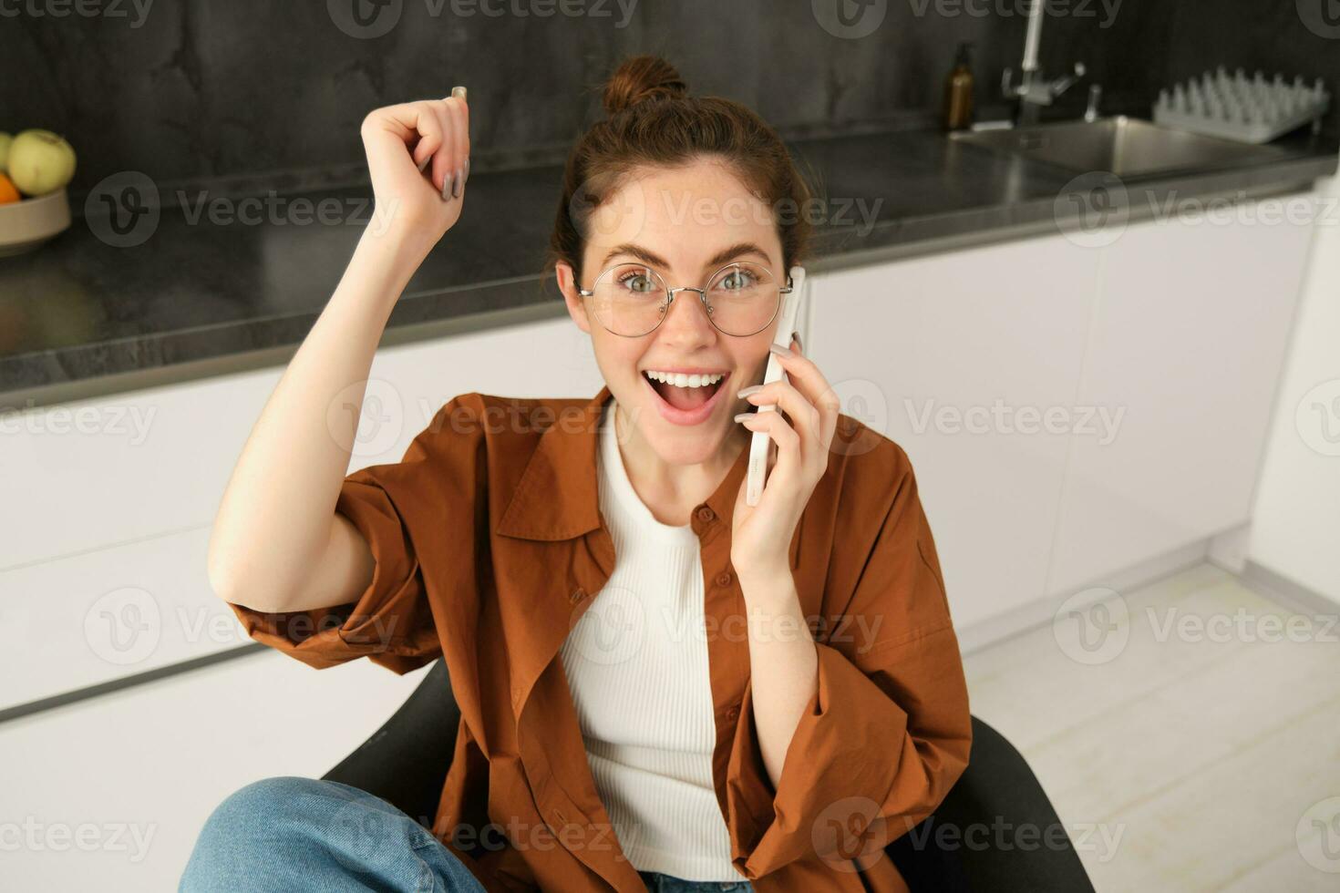Portrait of woman receiving great news over the phone. Girl talking on mobile telephone and celebrating, laughing and making fist pump, dancing on her chair in kitchen photo