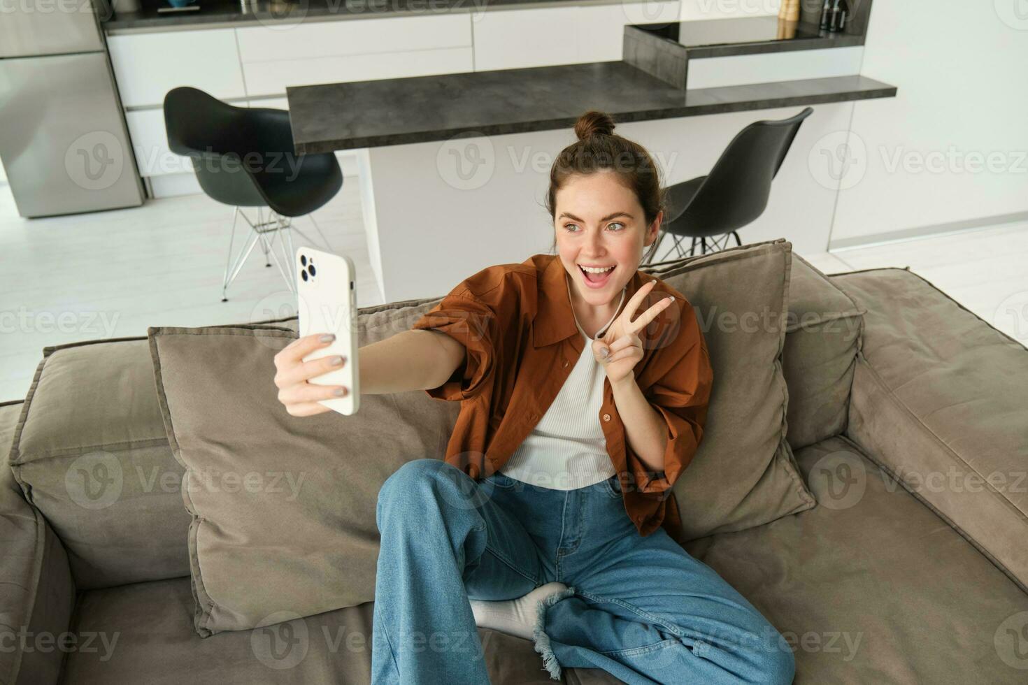 Portrait of cute smiling female model, takes selfie on smartphone app, posting photos on social media, using mobile phone camera, posing on sofa at home