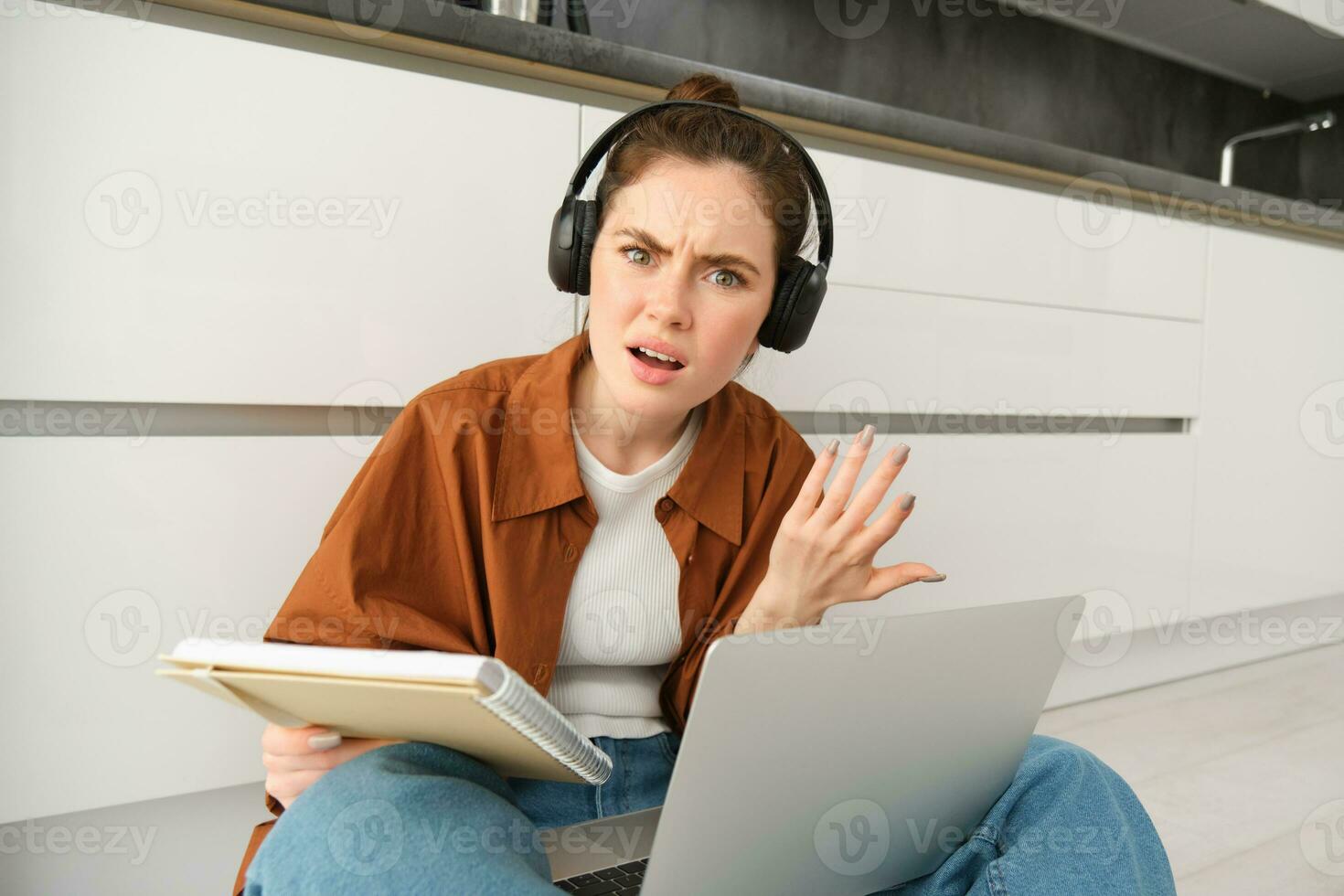 Portrait of annoyed and confused woman in headphones, sits on floor with laptop and notebook, looks frustrated, has issues in doing homework or studying photo