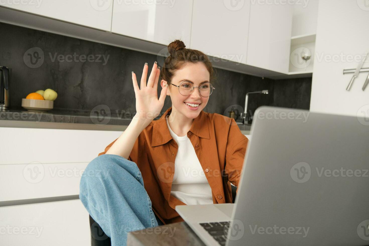 alegre joven mujer en anteojos, diciendo Hola, ondulación mano a computadora portátil, hablando a amigo en línea, vídeo charlando, sentado en cocina con computadora foto