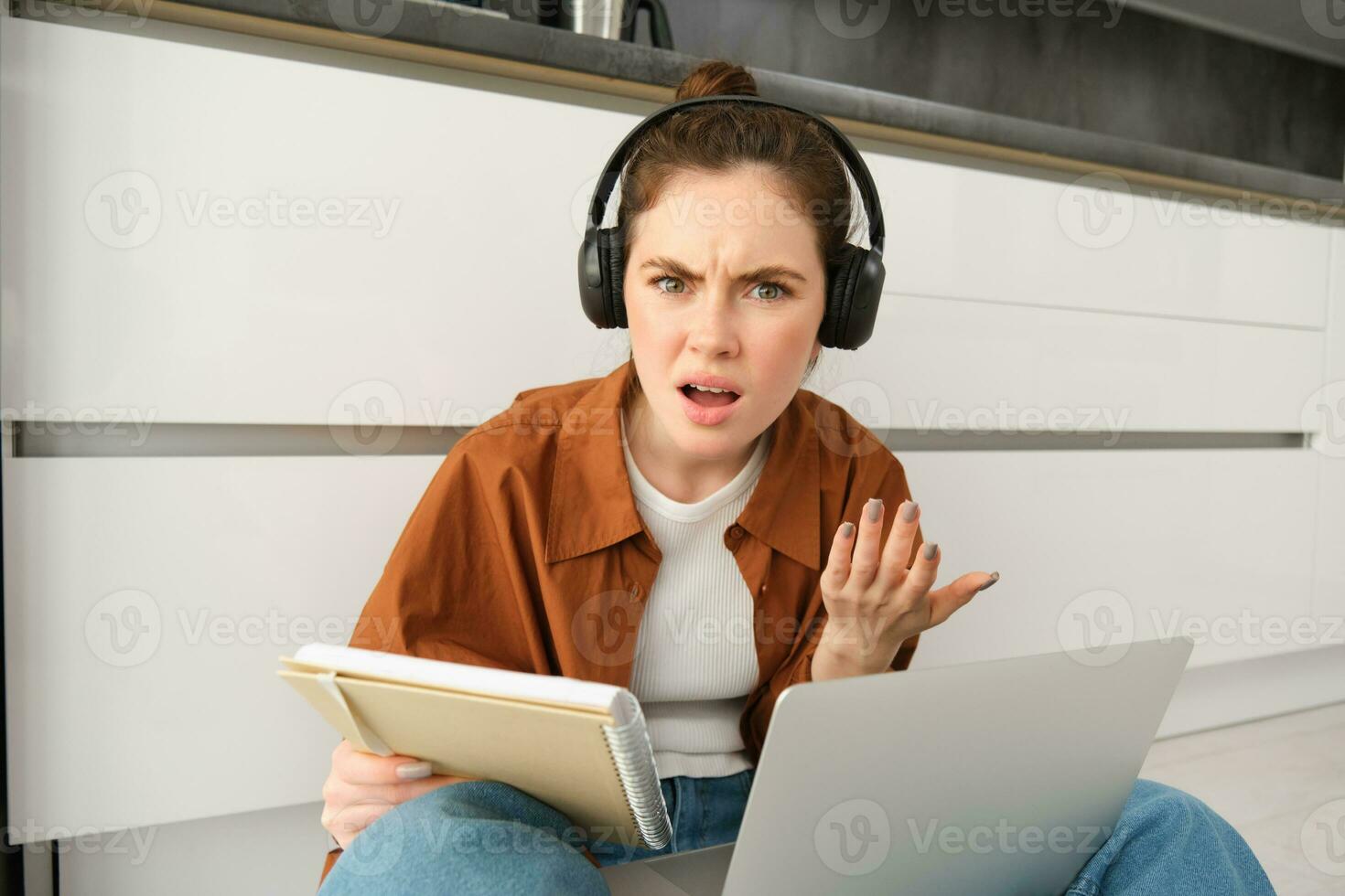Portrait of annoyed and confused woman in headphones, sits on floor with laptop and notebook, looks frustrated, has issues in doing homework or studying photo