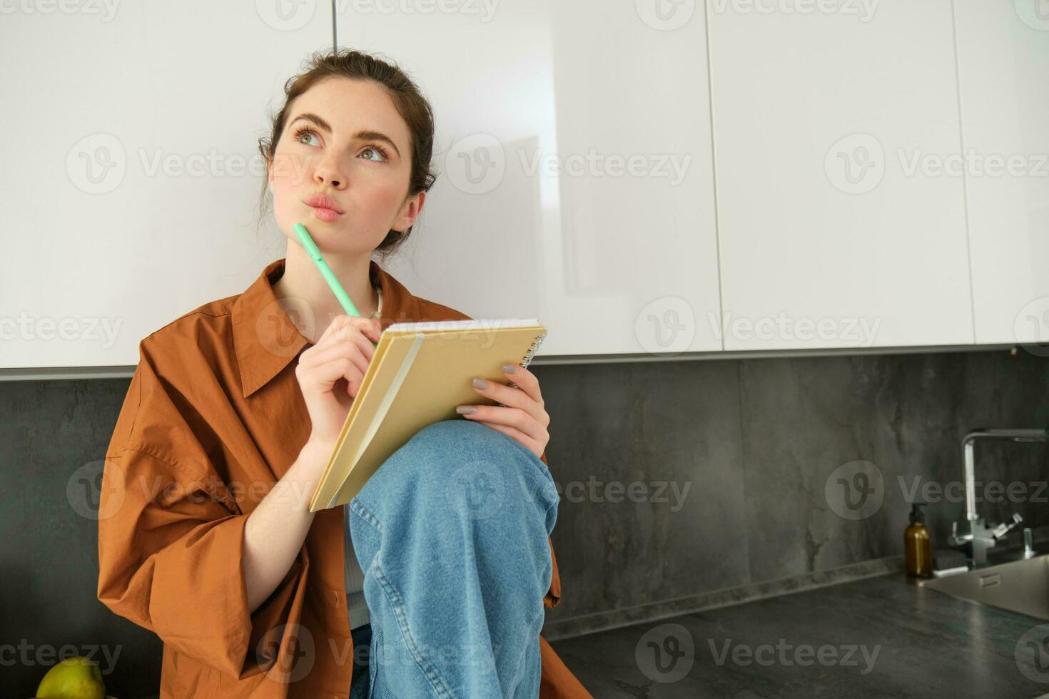 Image of young creative woman, artist drawing sketches in her notebook, sitting on kitchen counter at home with thoughtful face photo