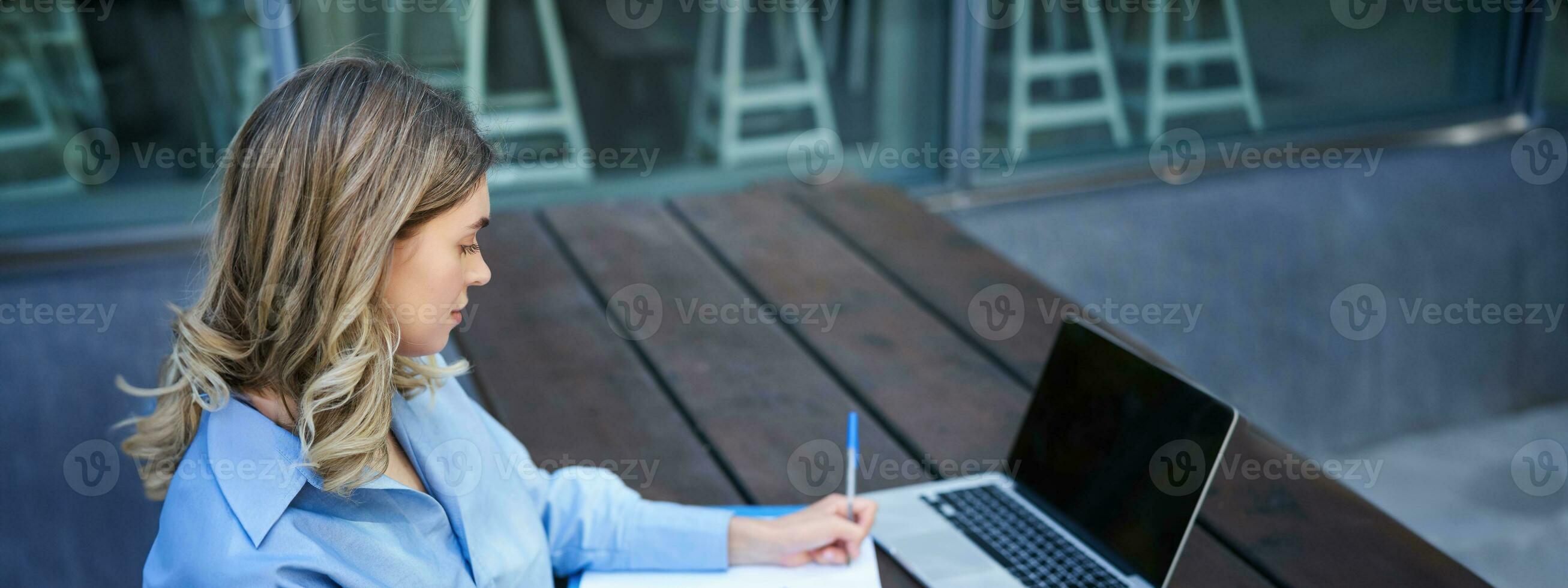 Portrait of businesswoman taking notes, studying on her computer, sitting outside in park. Young woman writing down, video conferencing on laptop photo