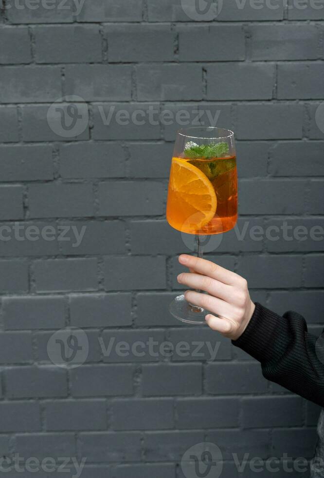 Close-up of woman's hand holding glass of cocktail with orange slice on black brick wall background photo