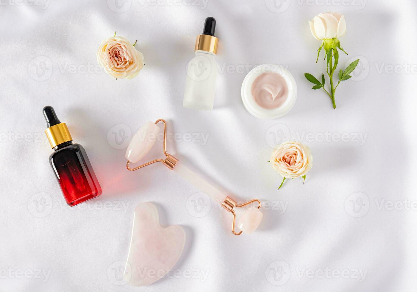 Top view of cosmetics in bottles and body care jar and quartz massager with gua sha scraper. Fabric white satin background. Flat lay. photo