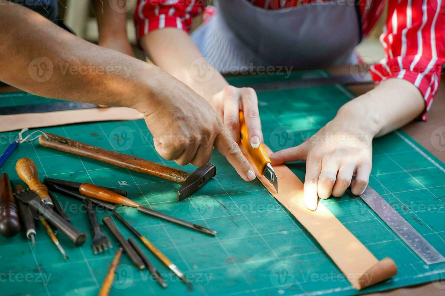 de cerca y cosecha mano de mayor artesano enseñando joven hembra cuero fabricante utilizando cortador cuchillo cortar un cueros a hacer cinturones en su trabajo tienda. foto