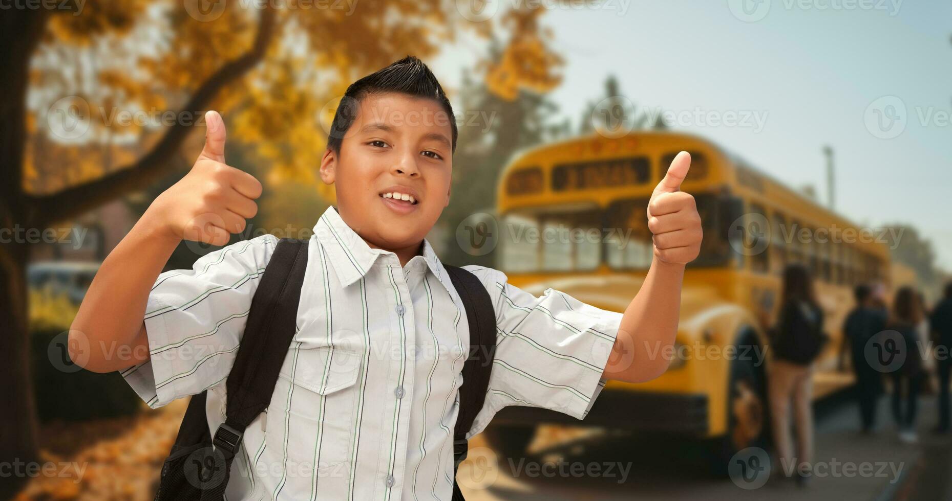 Excited Young Hispanic Boy Wearing a Backpack Giving Two Thumbs Up on Campus Near a School Bus photo