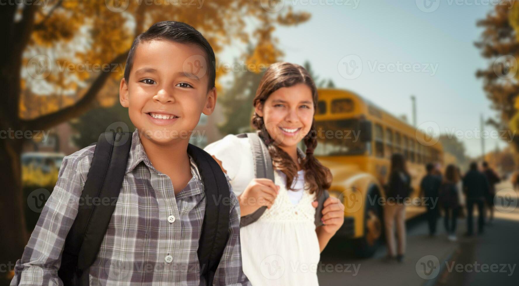 Happy Young Hispanic Boy and Girl Wearing Backpacks Near a School Bus on Campus photo