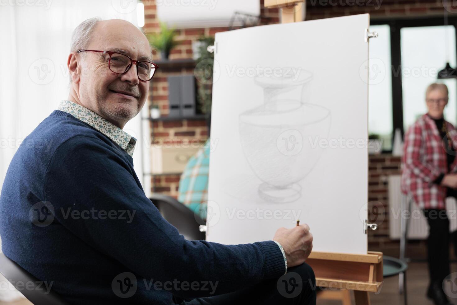 Happy mature man smiling at camera while drawing with pencil on canvas, senior people learning new skill, enjoying creative activities in retirement, taking up art activity at local community center photo