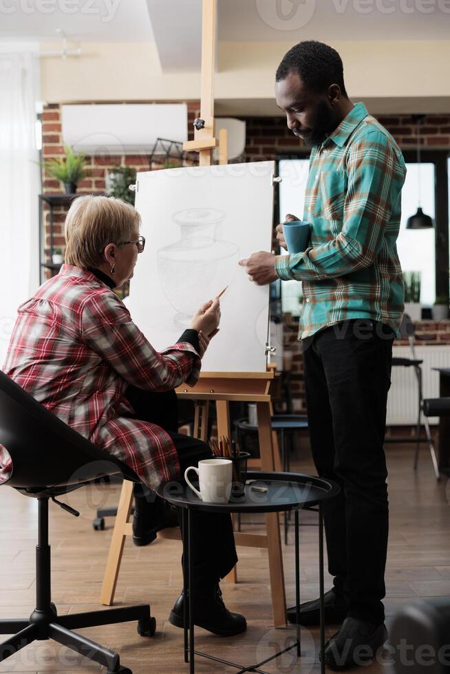 African American guy art instructor teaching senior woman to draw in class, standing near easel and explaining sketching techniques. Teacher assisting mature student during drawing lesson photo