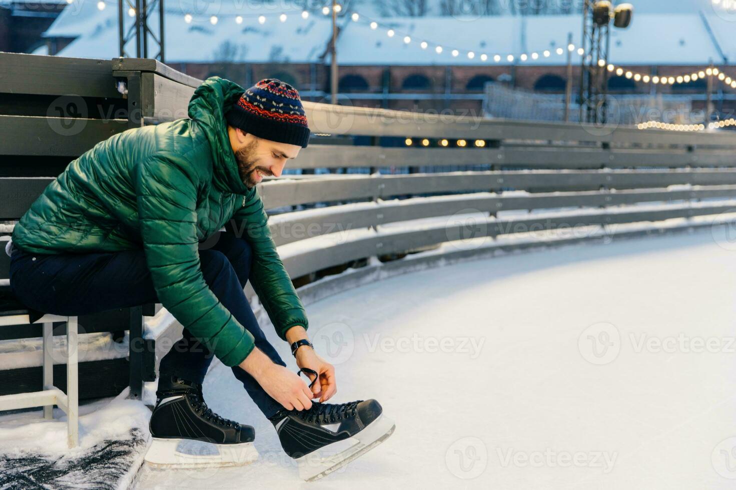 Focused man lacing up ice skates at a snowy rink, with warm lights in background photo