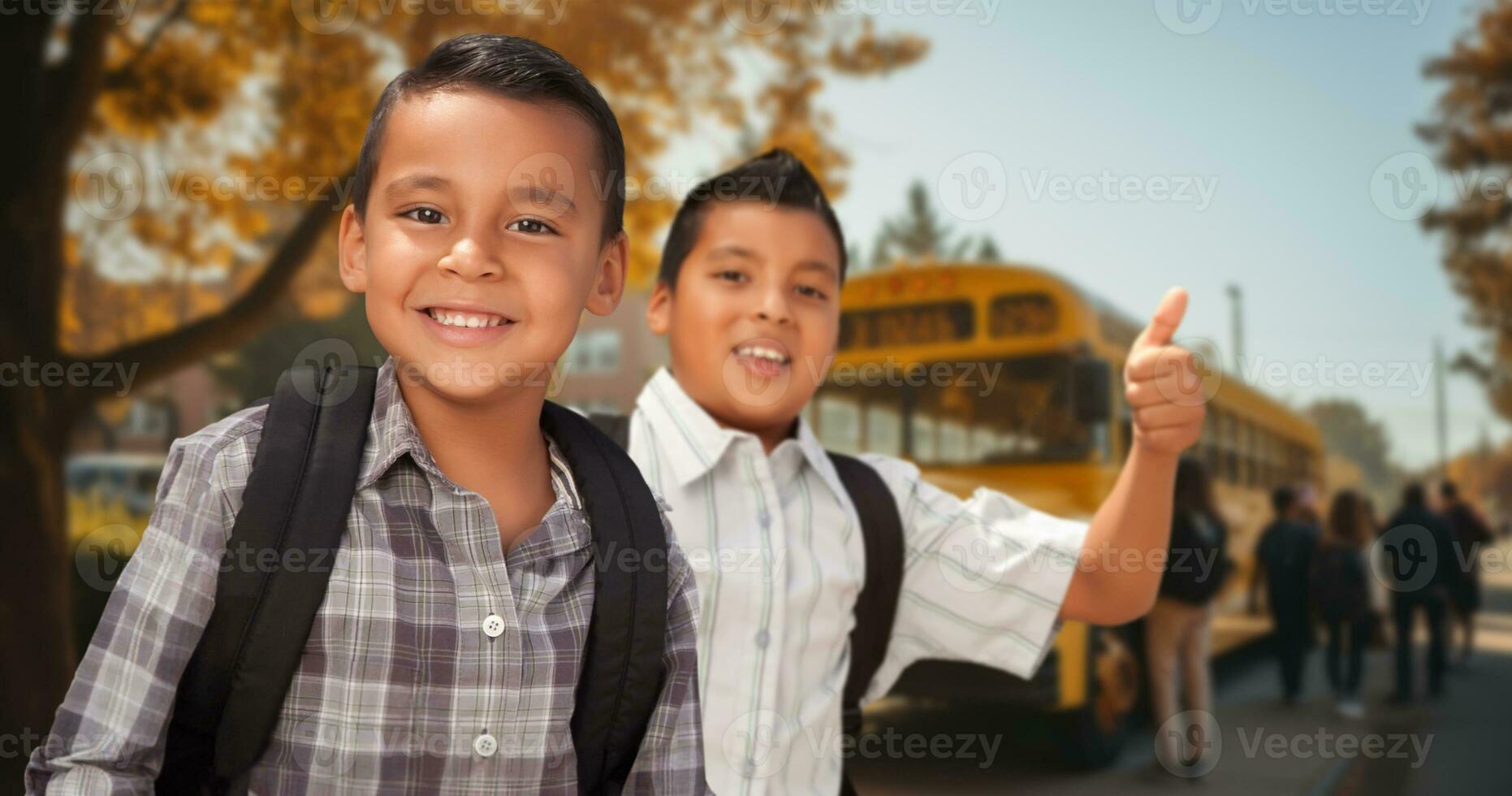 Two Happy Young Hispanic Boys Wearing Backpacks Give a Thumbs Up on Campus Near a School Bus photo