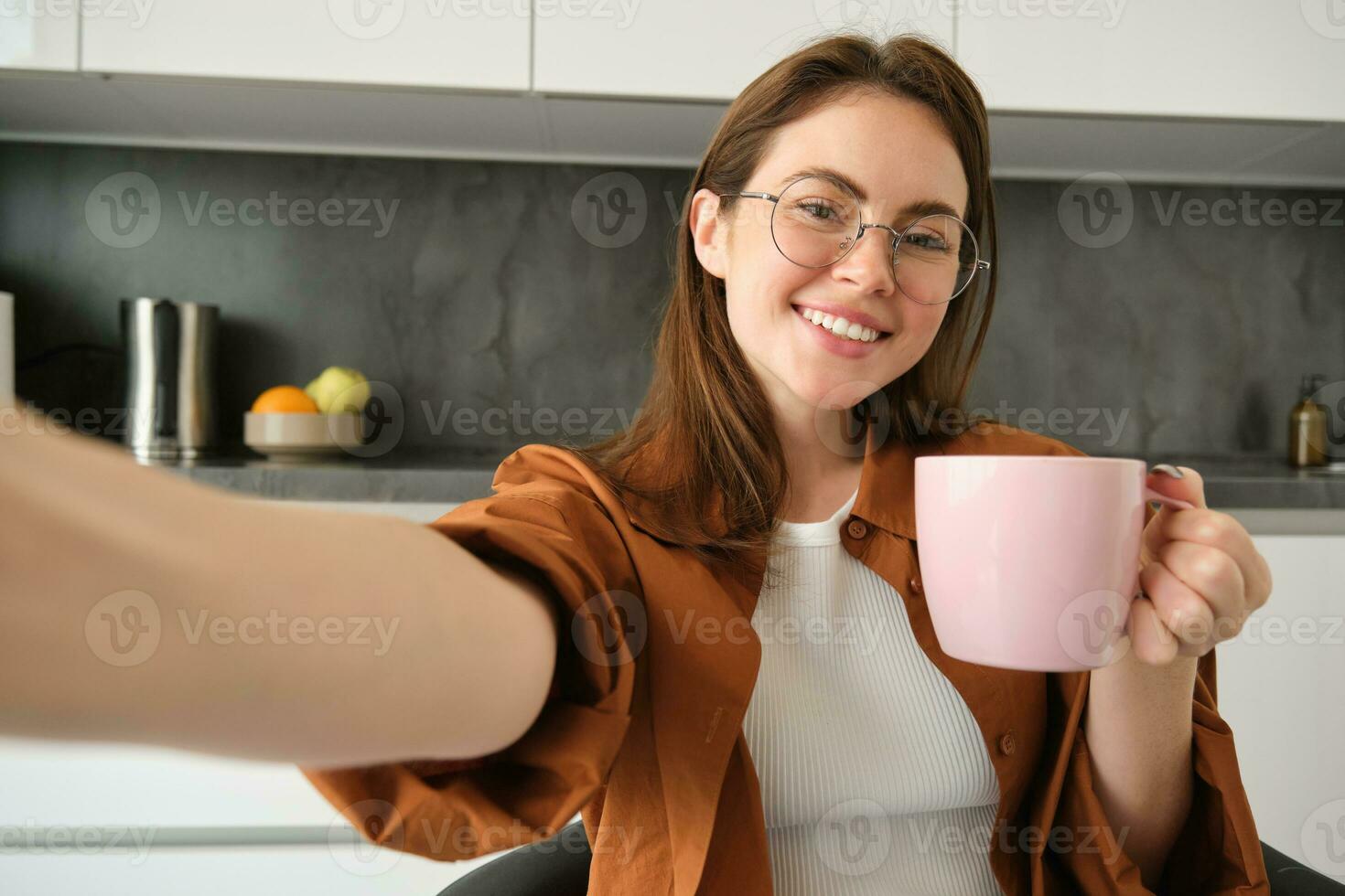 retrato de hermosa joven mujer en anteojos, tomando selfie en cocina con taza de Mañana café, haciendo fotos para su diario rutina estilo de vida Blog