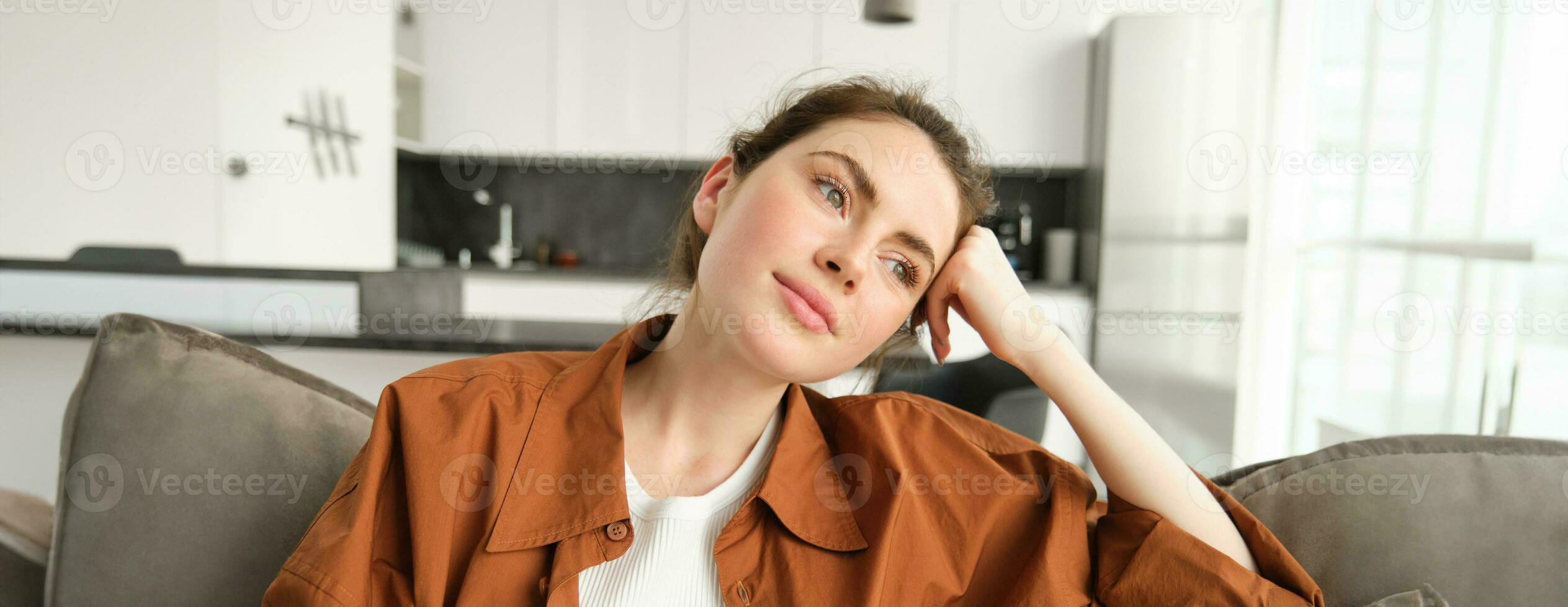 Lifestyle and people concept. Beautiful young brunette woman, sitting on couch with dreamy face, relaxing on sofa in living room, looking outside the window photo