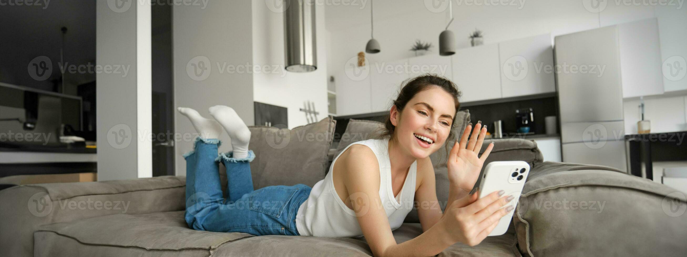 Happy young woman video chatting with friends, using smartphone app, waving hand at smartphone screen and saying hello photo