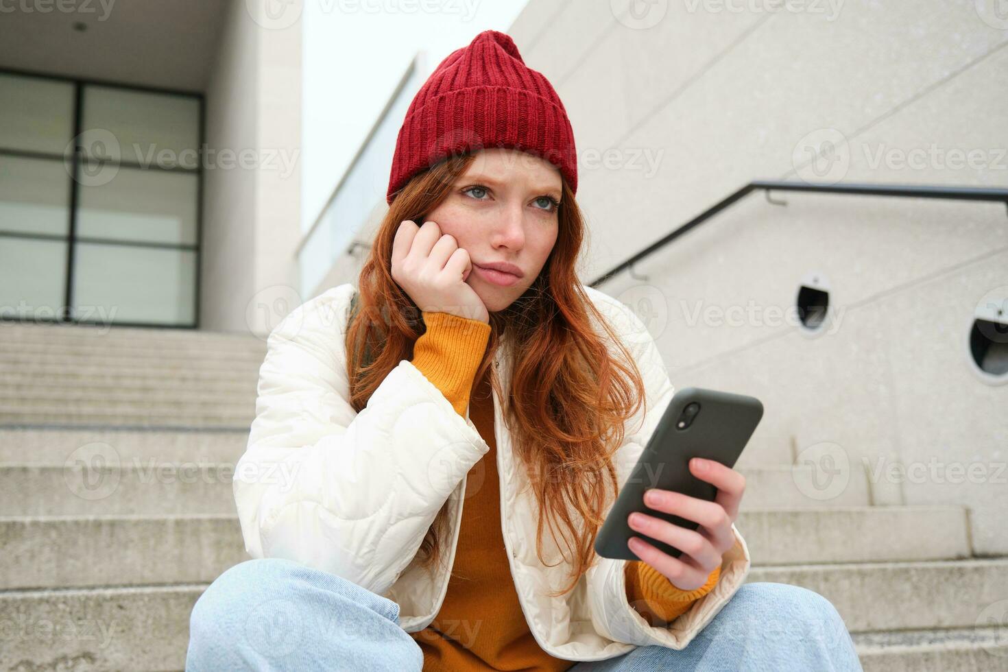 retrato de triste pelirrojo chica, mira trastornado y decepcionado a teléfono inteligente pantalla, lee malo Noticias texto mensaje en móvil teléfono y frunce el ceño, se sienta en escalera al aire libre foto