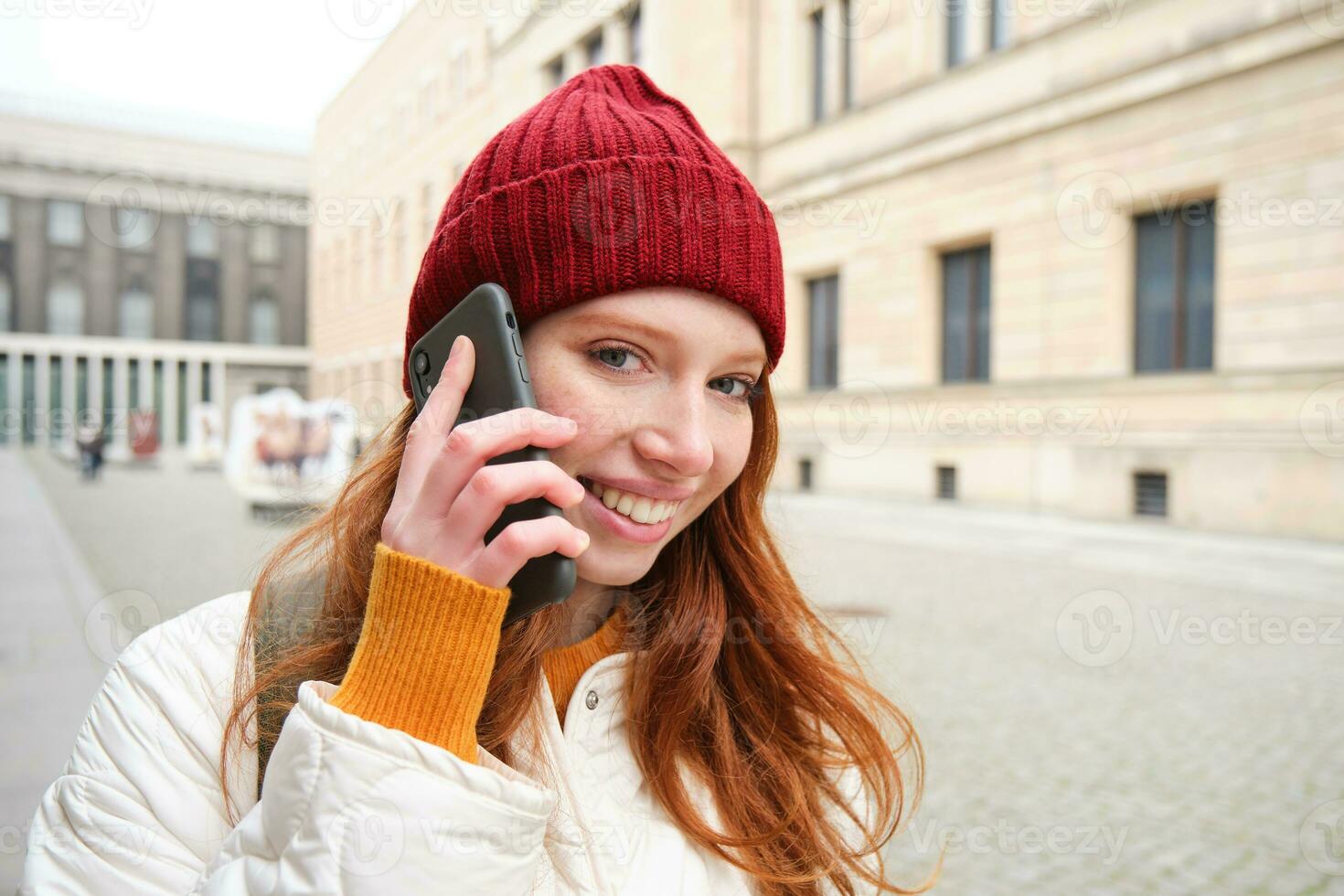 móvil banda ancha y gente. sonriente joven pelirrojo mujer camina en pueblo y negociaciones en móvil teléfono, vocación amigo en teléfono inteligente, utilizando Internet a hacer un llamada extranjero foto