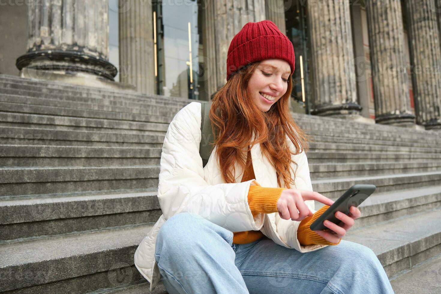 Portrait of young urban girl in red hat, sits on stairs near museum, holds mobile phone, connects to public wifi and surfs net, uses smartphone apps photo