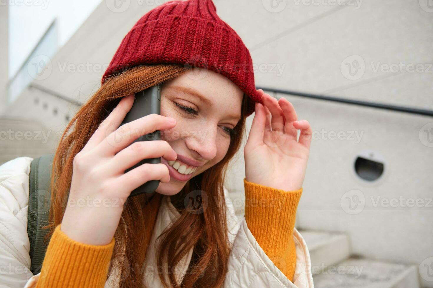 joven elegante pelirrojo niña en rojo sombrero, se sienta en calle y negociaciones en móvil teléfono, tiene teléfono conversación, anillos su amigo mientras relaja al aire libre foto