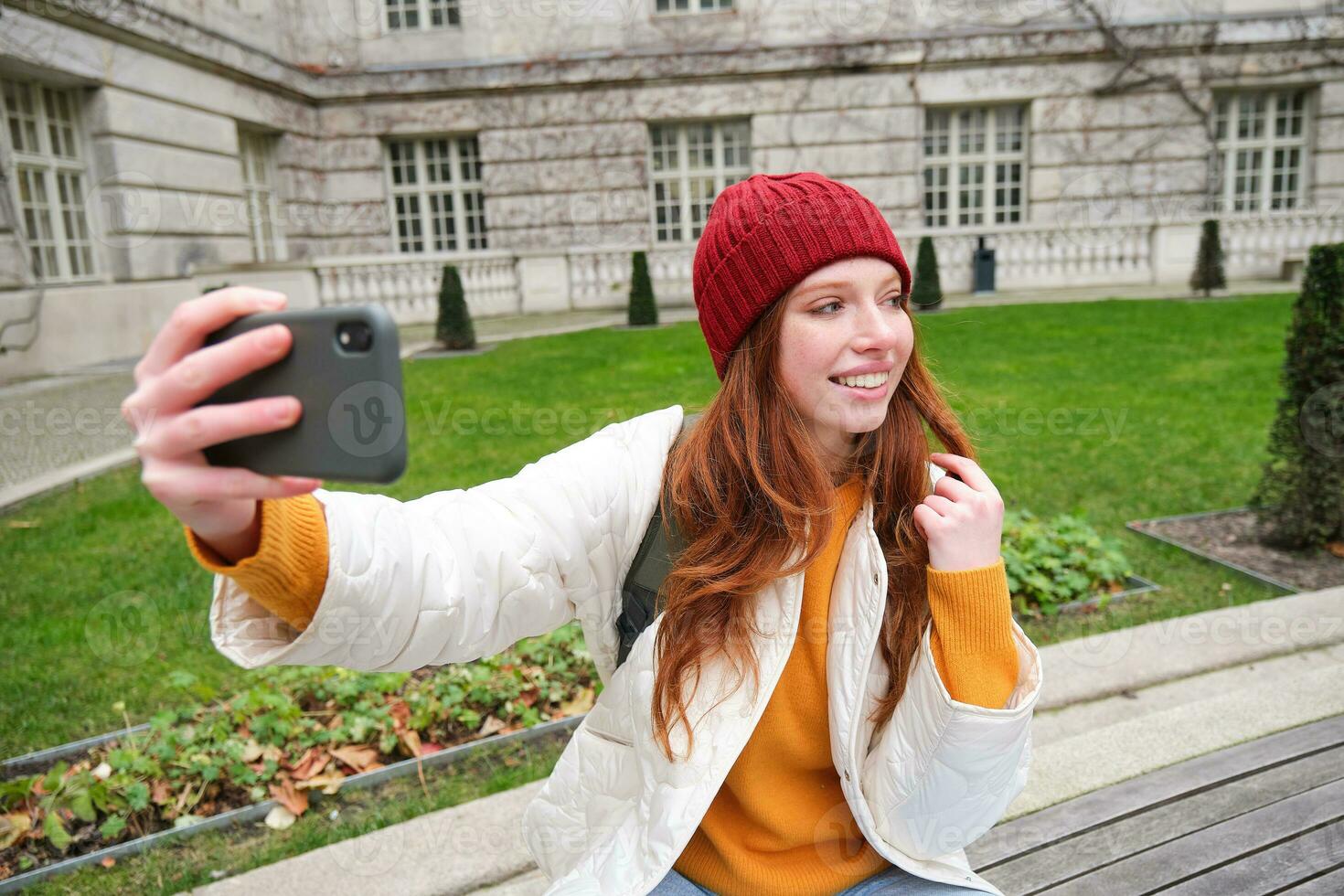 joven adolescente pelirrojo niña se sienta en banco en parque y toma autofoto, hace un foto de sí misma con teléfono inteligente aplicación, registros vlog