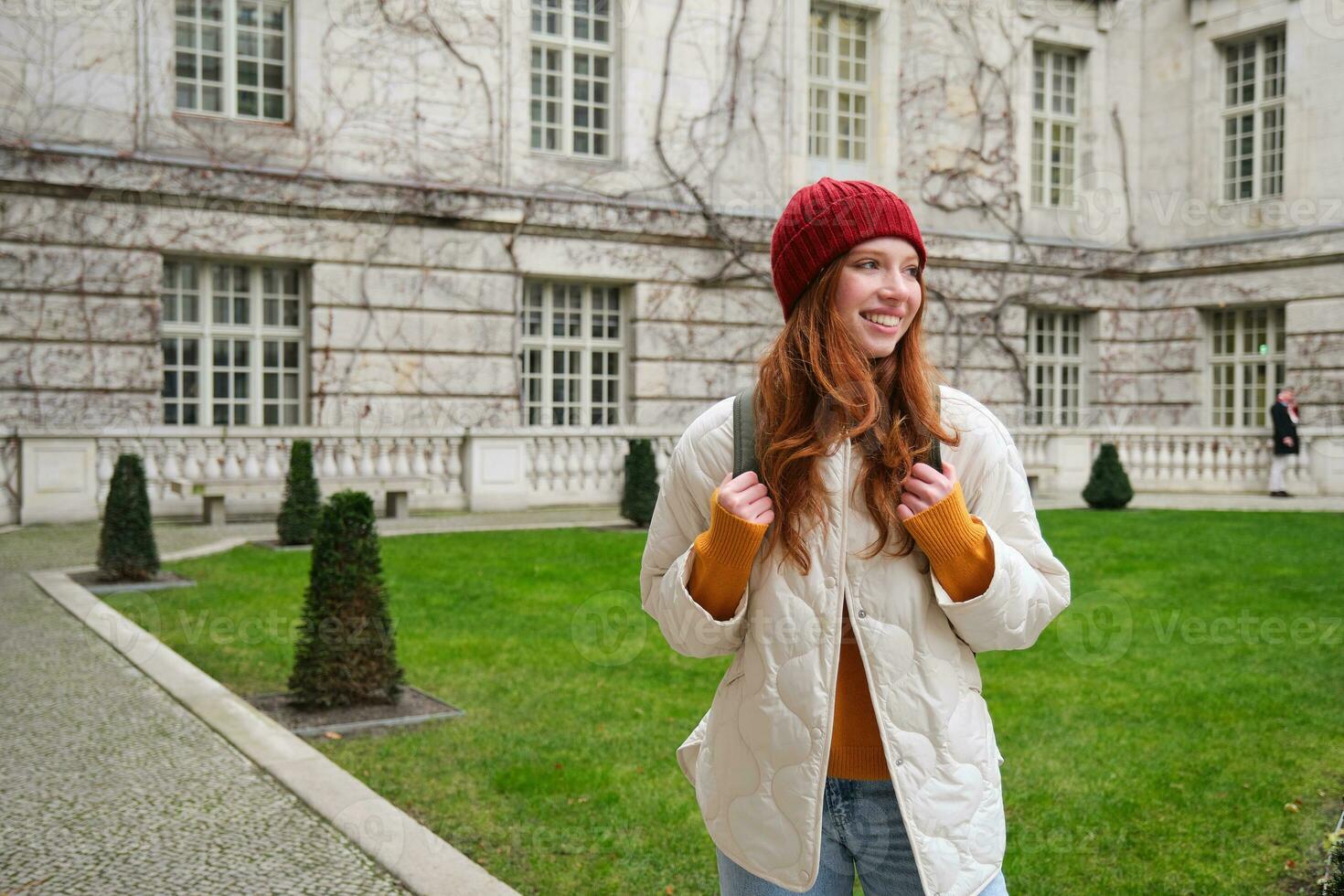 Young smiling redhead woman walking in beautiful city attractions, wearing backpack, red hat and coat, looking around with happy face photo