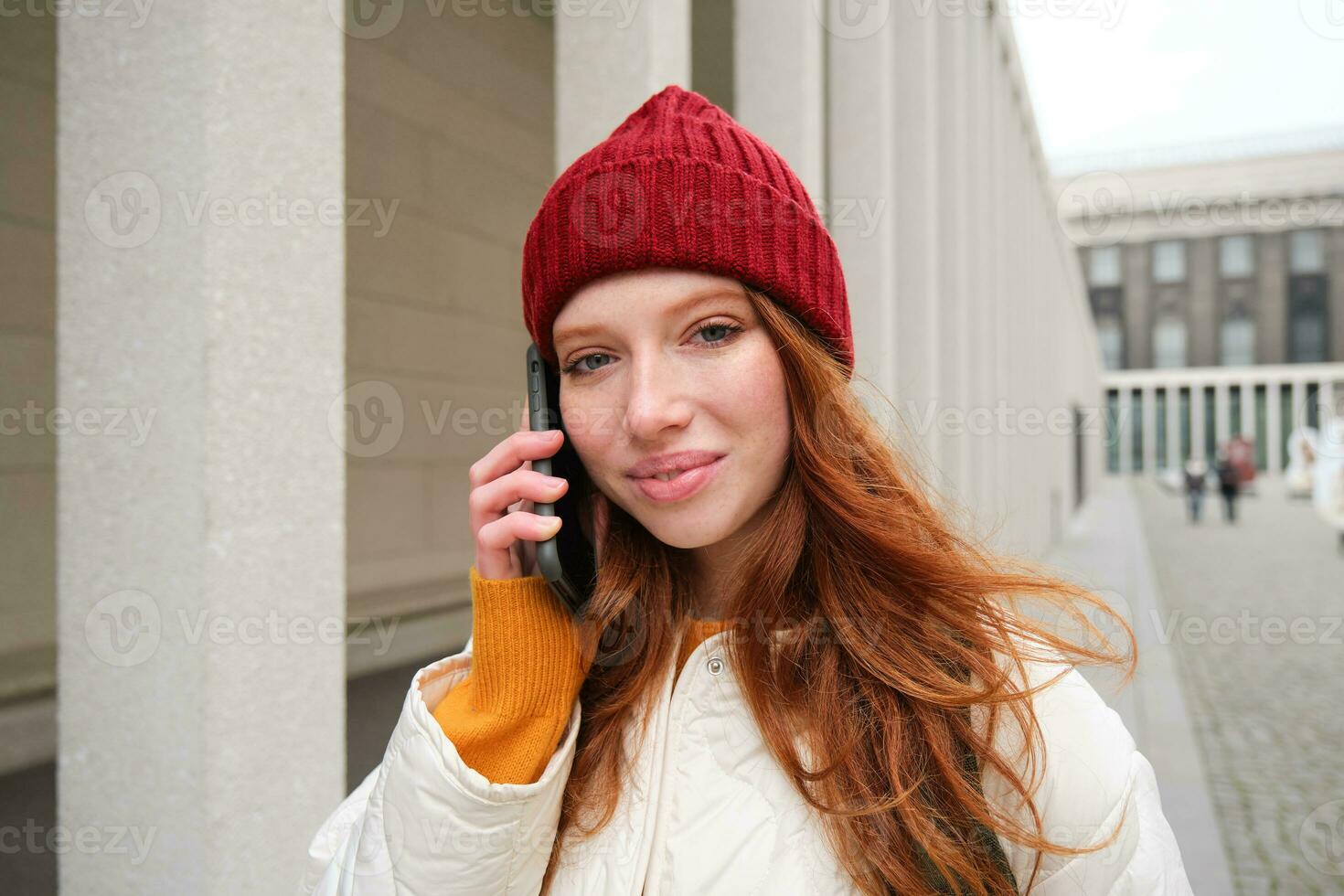 Mobile broadband and people. Smiling young redhead woman walks in town and talks on mobile phone, calling friend on smartphone, using internet to make a call abroad photo