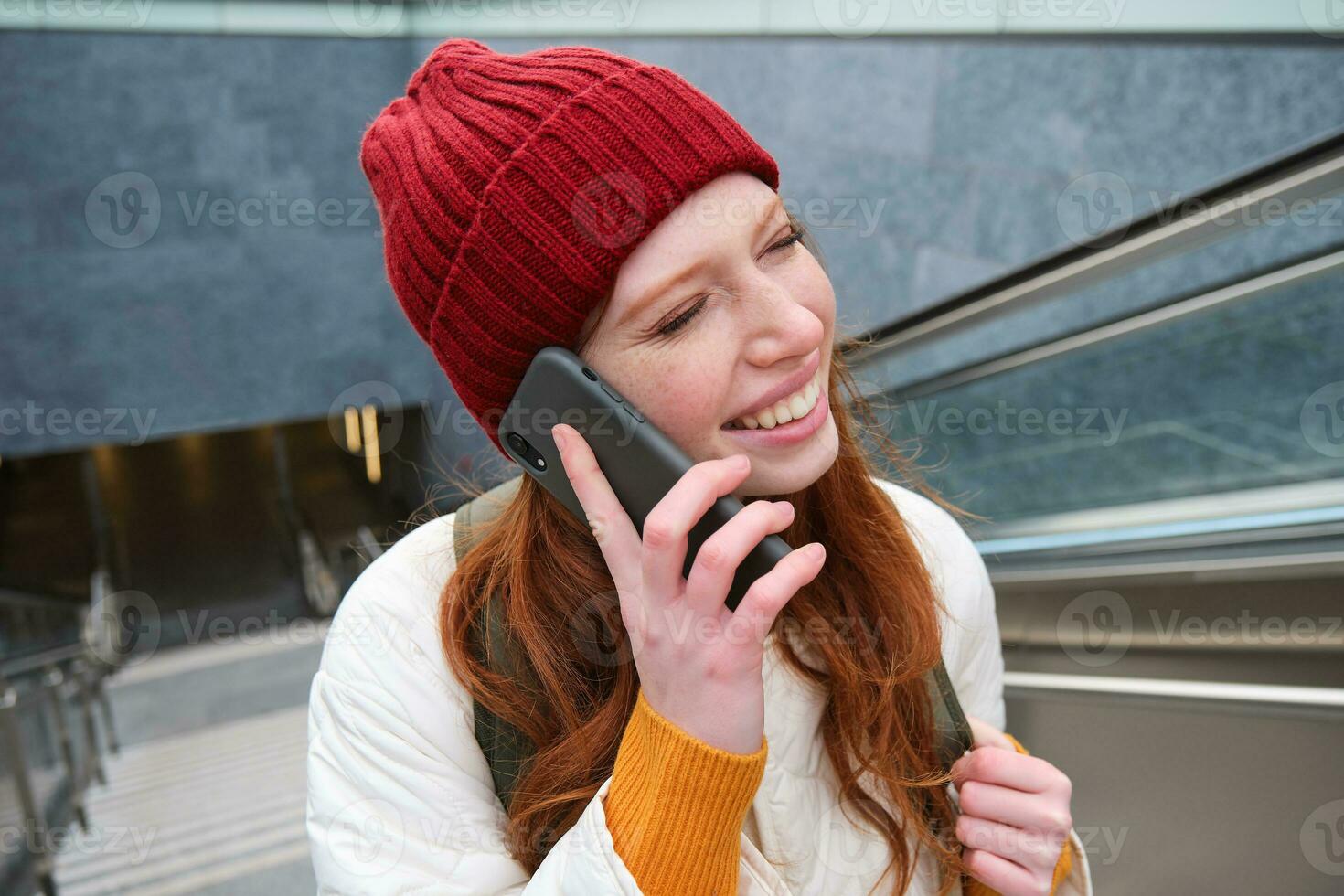 retrato de contento pelirrojo mujer caminando alrededor pueblo con teléfono inteligente, vocación alguien, hablando en móvil teléfono al aire libre foto