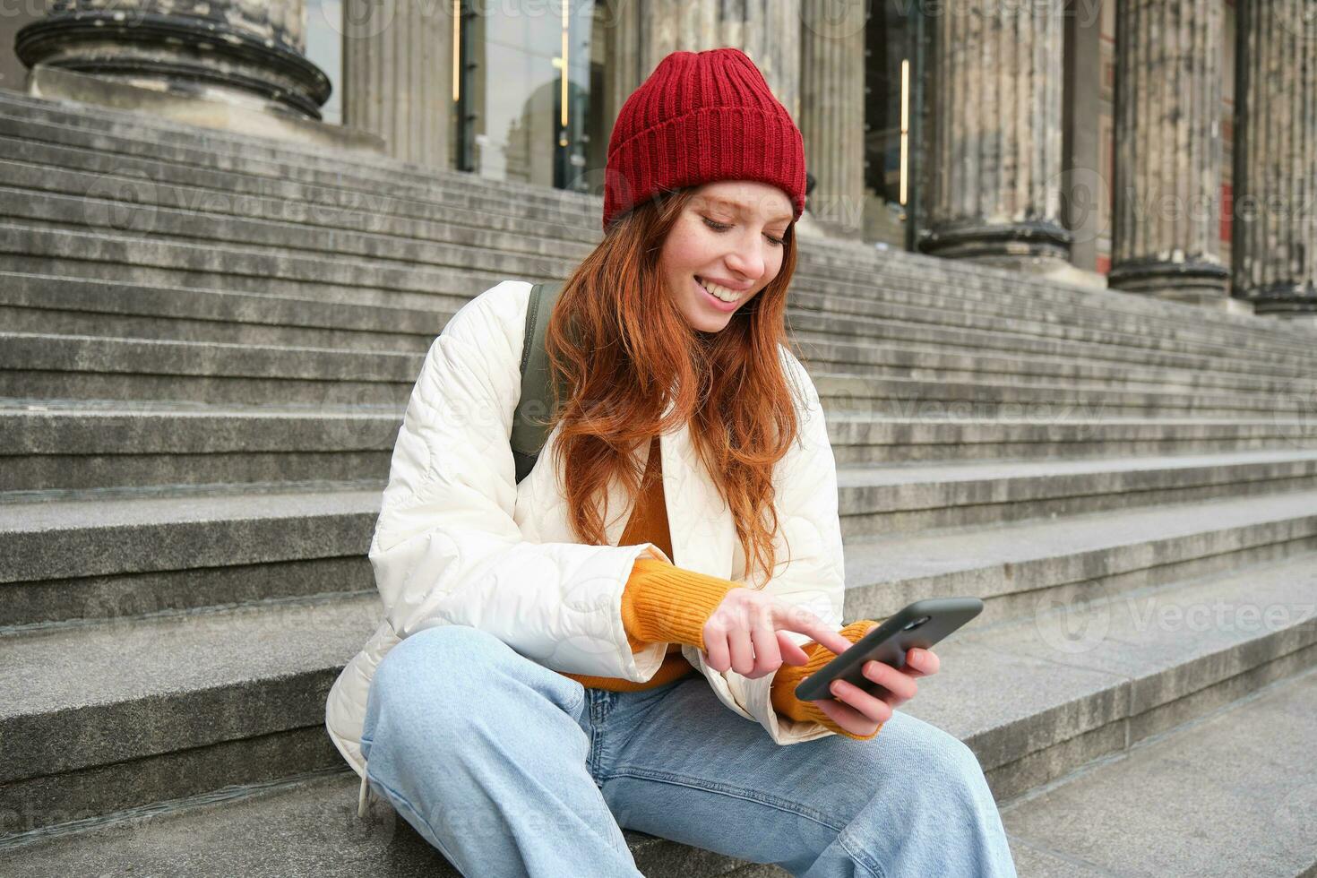 Portrait of young urban girl in red hat, sits on stairs near museum, holds mobile phone, connects to public wifi and surfs net, uses smartphone apps photo