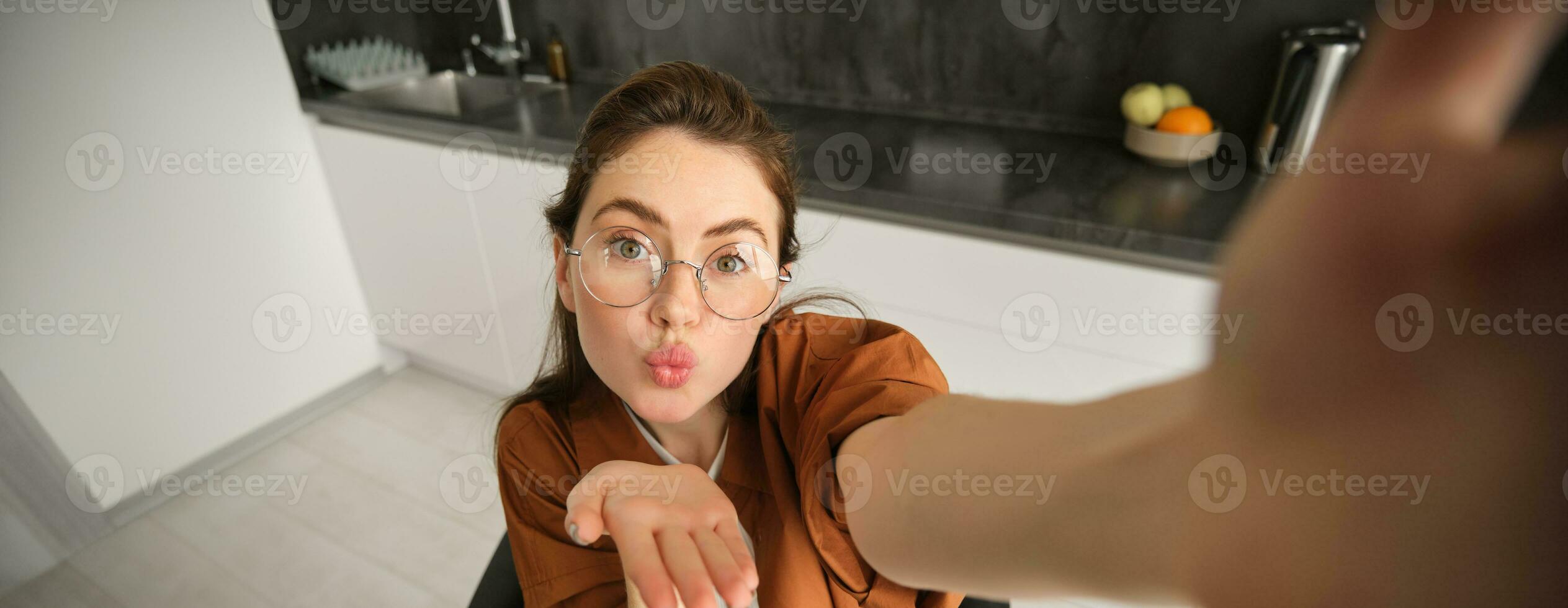 Selfie of happy young and carefree woman, taking photo on mobile phone with extended hand, posing and smiling, sitting in kitchen in glasses