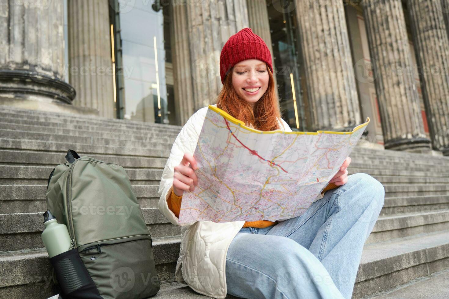 Portrait of young redhead woman, tourist sits with paper map and looks for a route to tourism attraction, rests on stairs outdoors photo