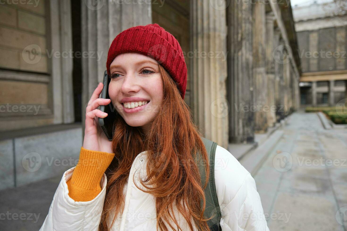 sonriente pelirrojo hembra turista negociaciones en móvil teléfono y camina alrededor ciudad. contento estudiante en rojo sombrero llamadas amigo, soportes en calle y usos teléfono inteligente foto