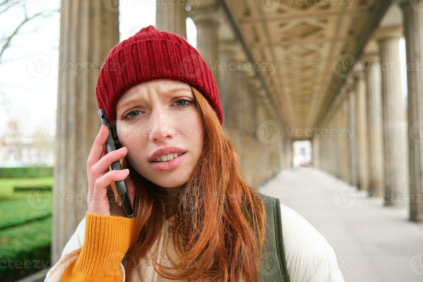 retrato de pelirrojo niña en rojo sombrero, llamadas alguien, escucha a voz mensaje con preocupado, confuso cara expresión, utilizando teléfono inteligente foto