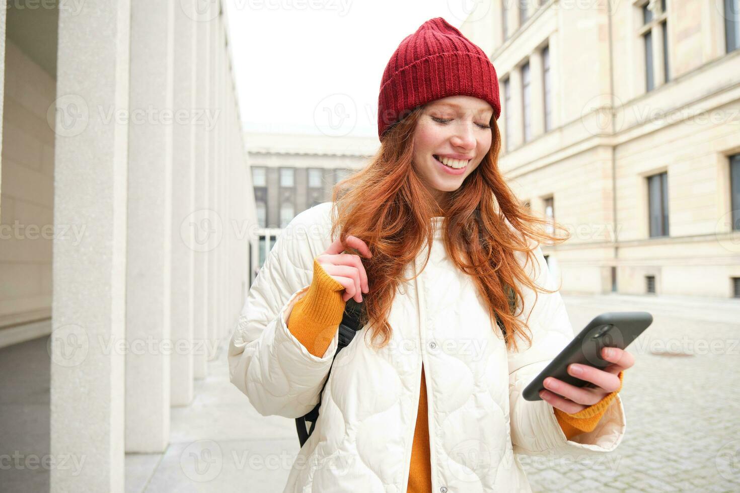 Beautiful smiling girl, tourist with backpack, holding smartphone, using map on mobile phone application, looking for sightseeing in internet app, standing outdoors photo