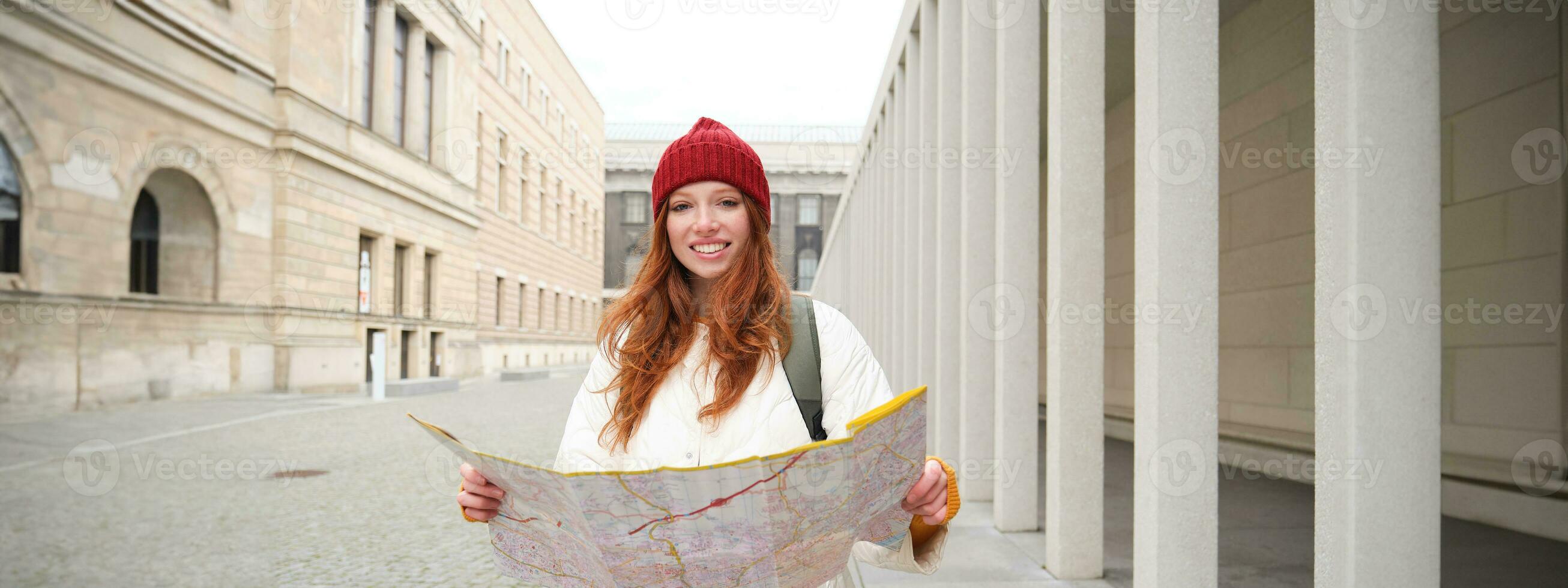 Beautiful redhead woman, tourist with city map, explores sightseeing historical landmark, walking around old town, smiling happily photo