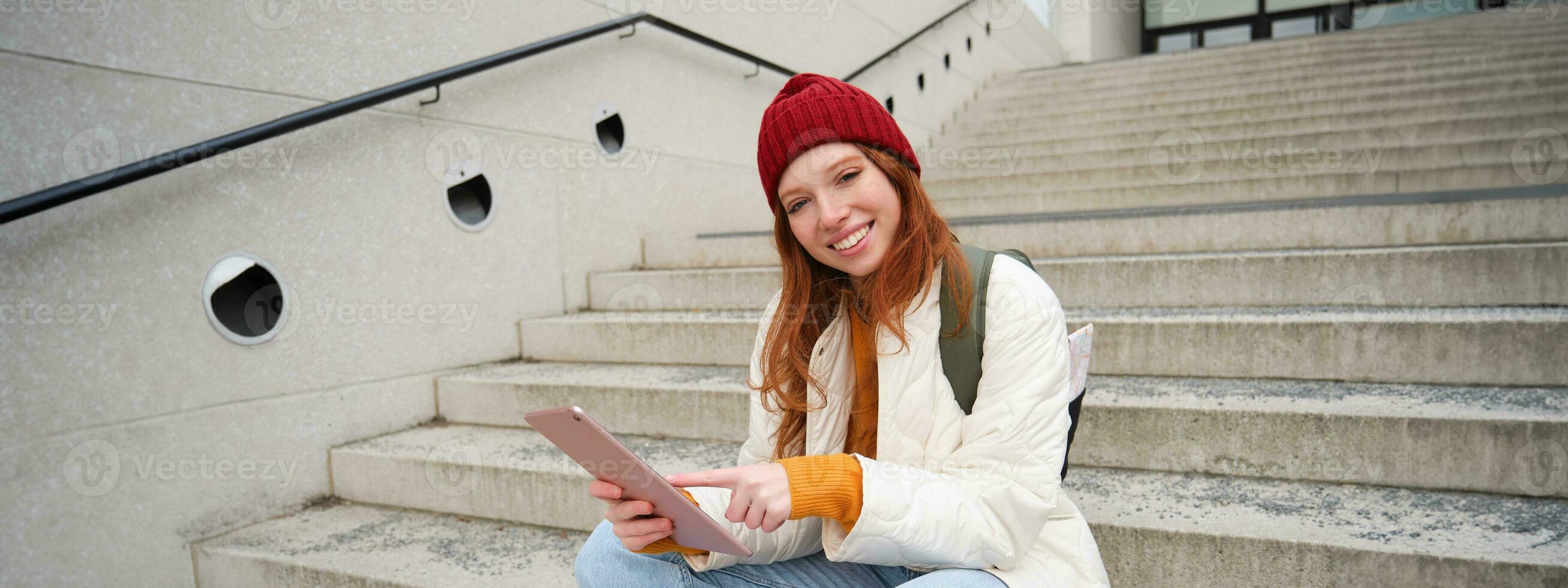 Happy stylish redhead girl, student in red hat, holds digital tablet, uses social media app, searches something online, connects to wifi photo
