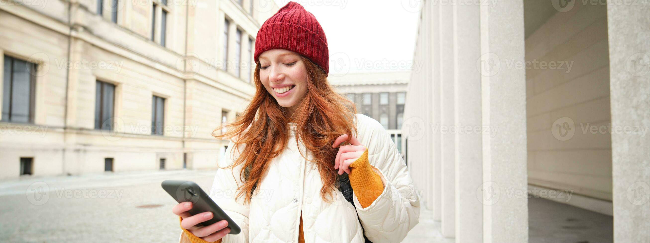 Beautiful smiling girl, tourist with backpack, holding smartphone, using map on mobile phone application, looking for sightseeing in internet app, standing outdoors photo