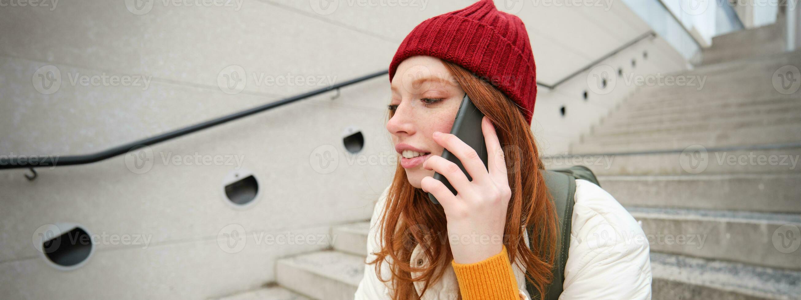joven elegante pelirrojo niña en rojo sombrero, se sienta en calle y negociaciones en móvil teléfono, tiene teléfono conversación, anillos su amigo mientras relaja al aire libre foto